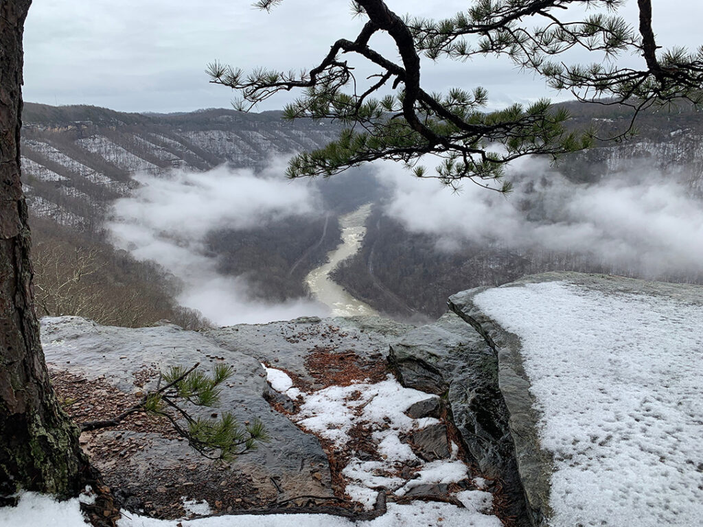 Winter view of New River Gorge National Park Endless Wall Trail snow covering rocks with a view of the New River with clouds and trees 
